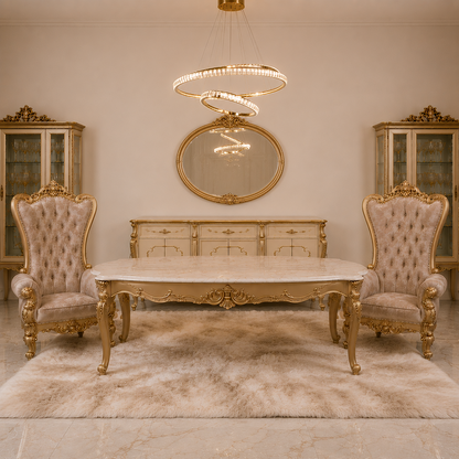 An ornate luxury dining room setup featuring a rectangular marble-top table with carved gold legs, flanked by two matching tufted wingback chairs, set on a plush cream rug. The background includes a gold-framed mirror, a cream sideboard, and matching glass-front cabinets under a crystal chandelier.