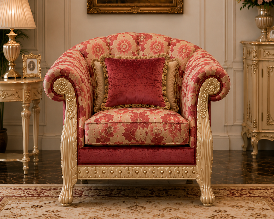 An ornate vintage-style armchair upholstered in a pink and beige floral patterned fabric, featuring carved cream-colored wood arms and legs, a matching red velvet accent cushion, and a coordinating red throw pillow, set in an elegant classical room.