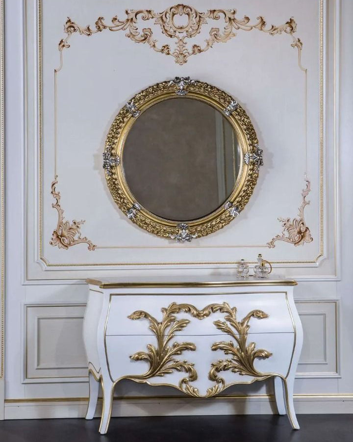 Decorative white cabinet with gold leaf detailing and a round mirror on a similarly decorated wall.